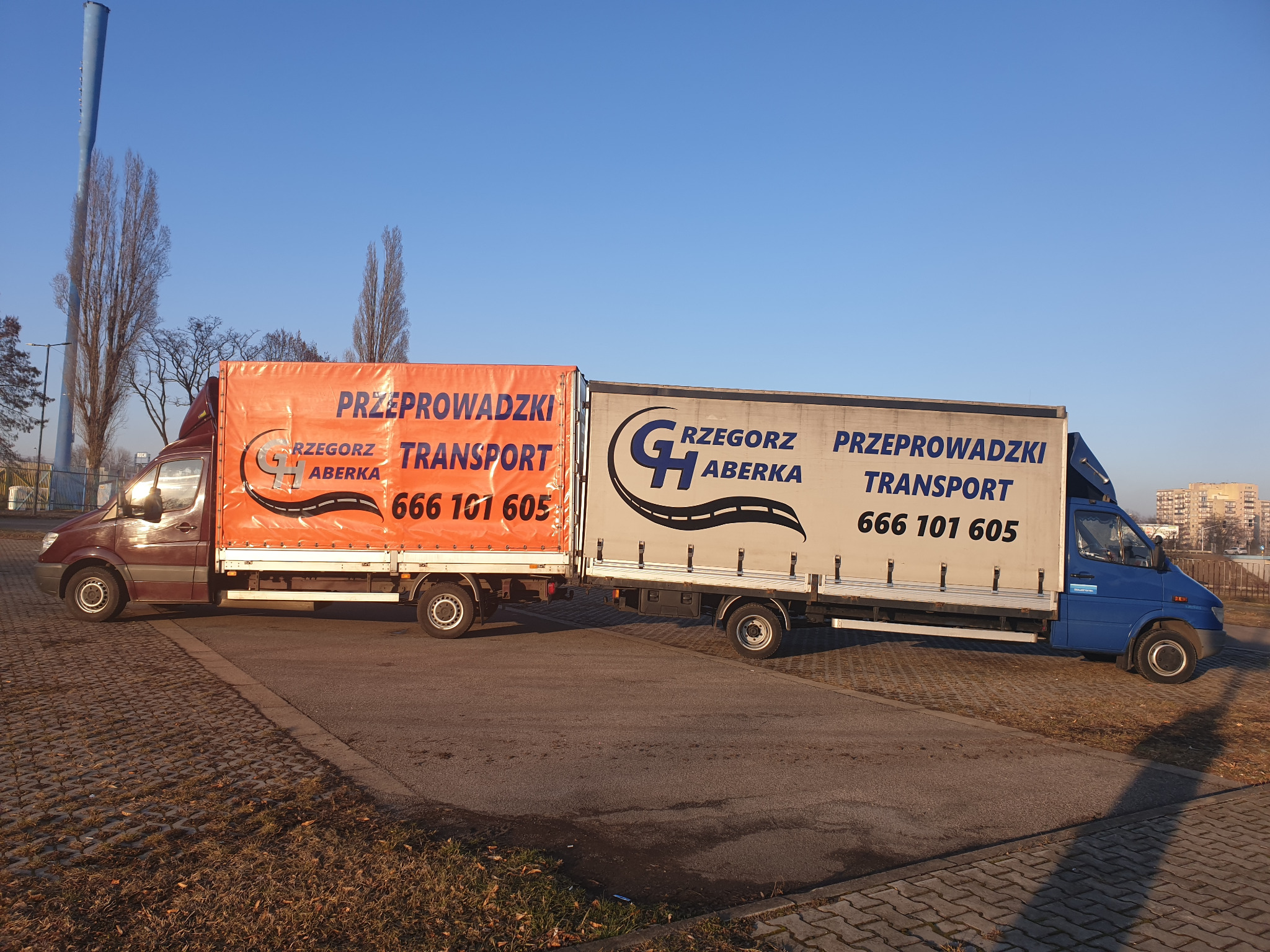 Dwa busy transportowe, jeden pomarańczowy, drugi niebieski, z napisem 'Przeprowadzki Transport' i numerem telefonu, stojące obok siebie na parkingu w słoneczny dzień.