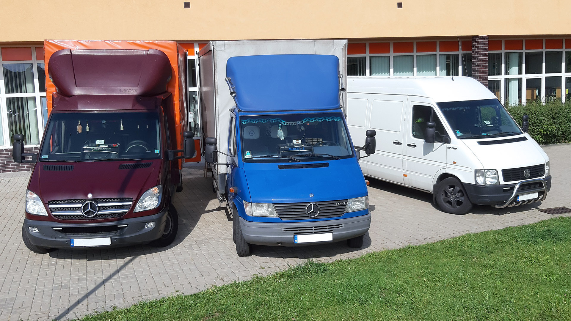 Trzy busy dostawcze Mercedes, zaparkowane obok budynku z pomarańczową fasadą, każdy w innym kolorze: bordowy, niebieski i biały, z widocznymi detalami jak lusterka i logo Mercedesa.