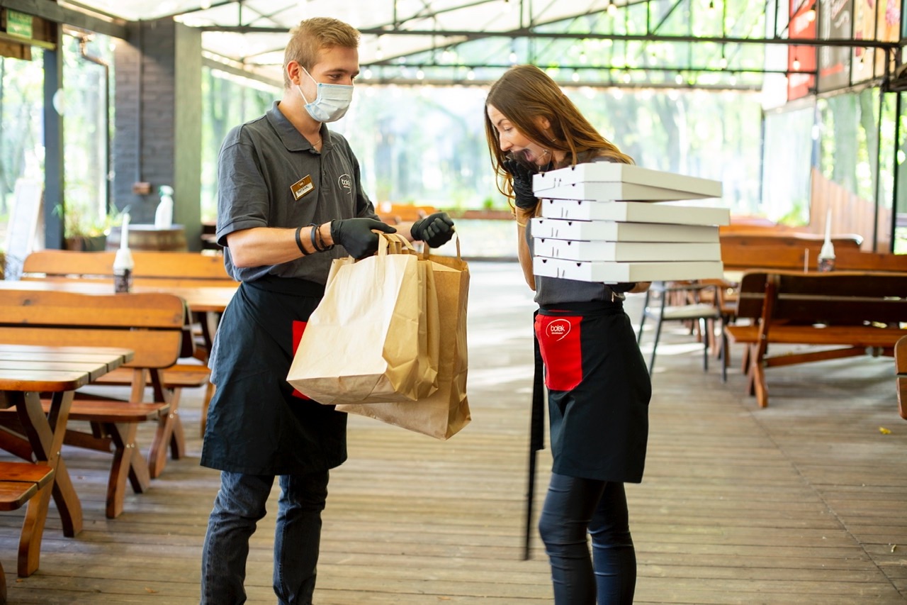 Kelner w maseczce i czarnych rękawiczkach przekazuje torby z zamówieniem kobiecie trzymającej stos pudełek na pizzę na tle drewnianych stołów i ławek.