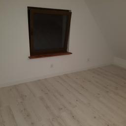 Ryszard Grudzień - Puste poddasze z drewnianą podłogą i oknem w brązowej ramie, widoczne gniazdka elektryczne na ścianie.