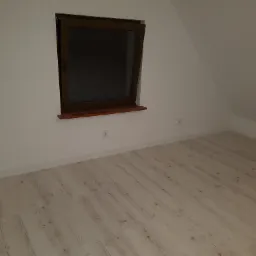 Puste poddasze z drewnianą podłogą i oknem w brązowej ramie, widoczne gniazdka elektryczne na ścianie.