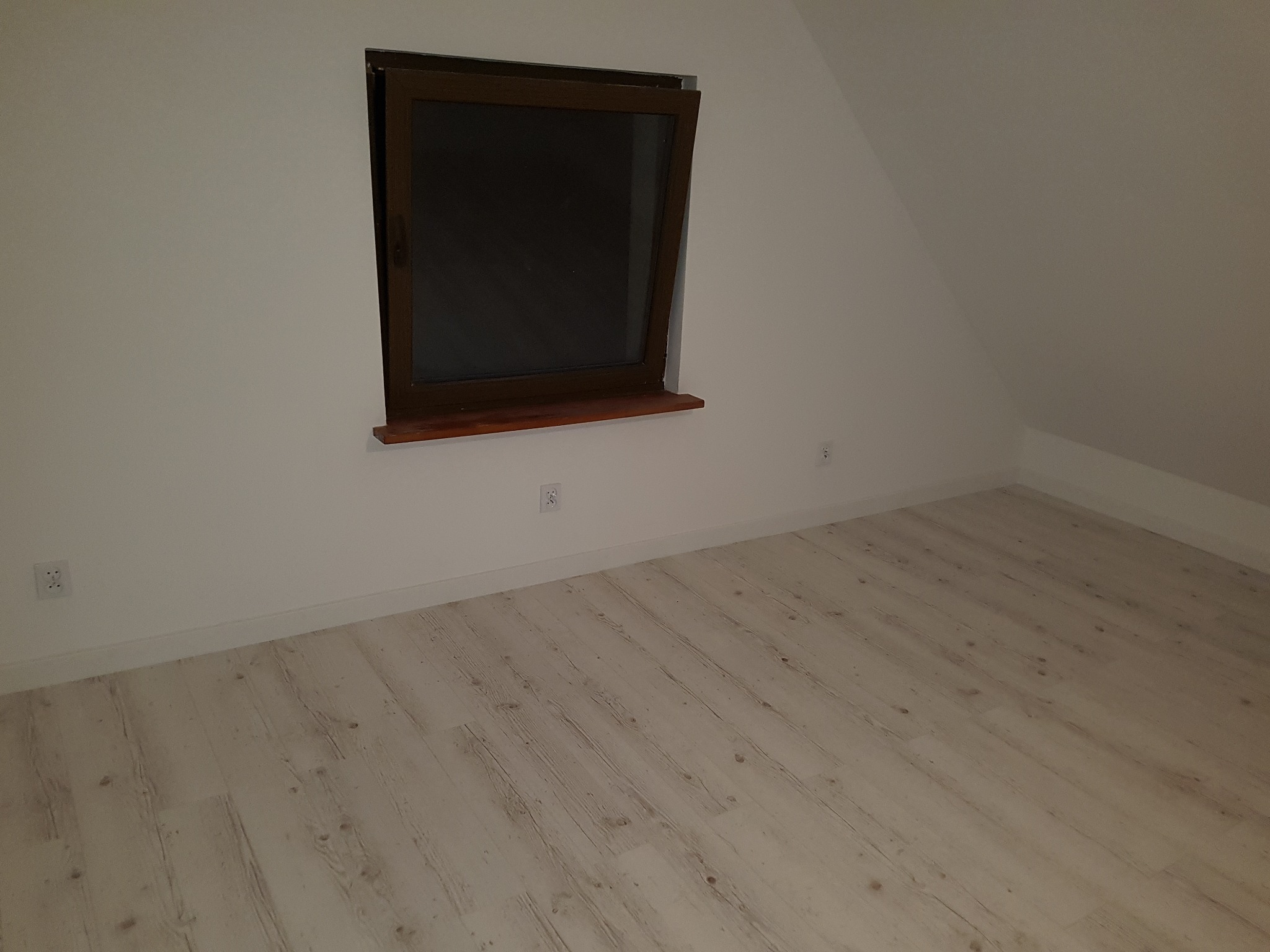 Puste poddasze z drewnianą podłogą i oknem w brązowej ramie, widoczne gniazdka elektryczne na ścianie.