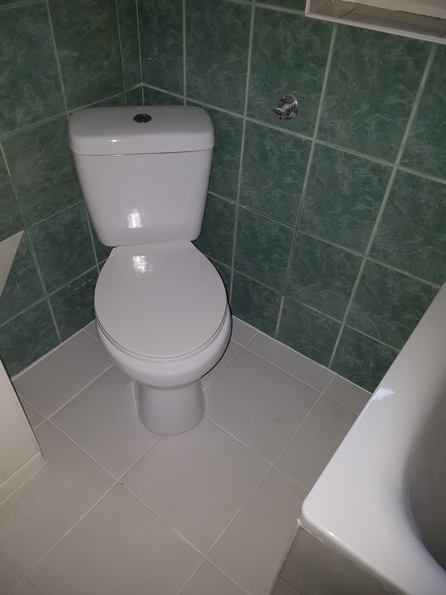 Biała toaleta kompaktowa po remoncie, widoczne zielone płytki ścienne z marmurkowym wzorem i szare płytki podłogowe, fragment wanny po prawej stronie.