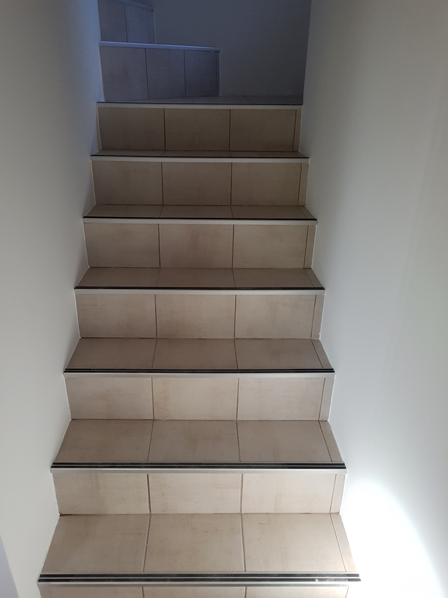 Schody wyłożone beżowymi płytkami ceramicznymi z czarnymi listwami antypoślizgowymi, widok z dołu na górę.
