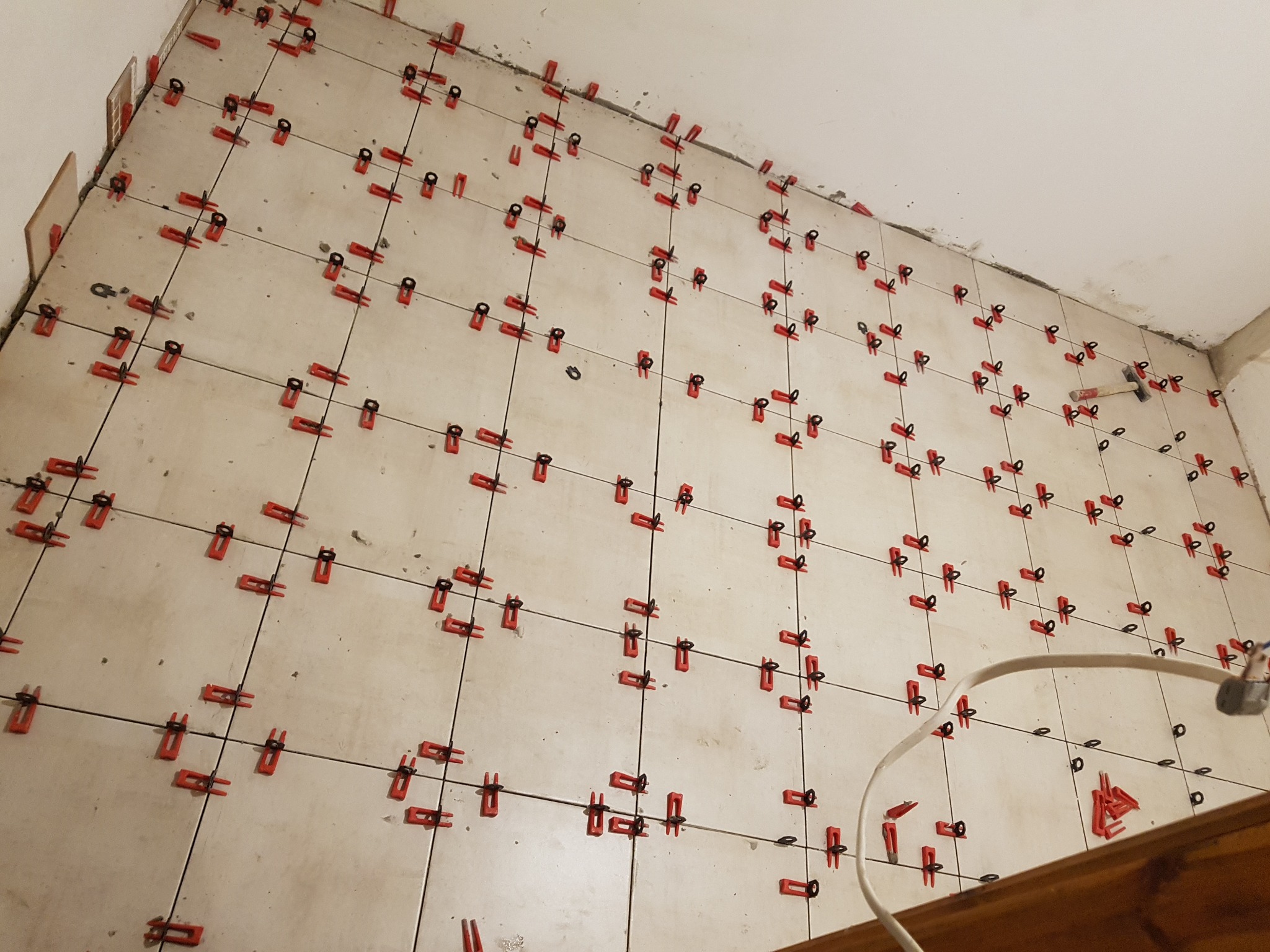 Układanie jasnych płytek na ścianie, system poziomowania z czerwonymi klipsami i czarnymi klinami, widoczny młotek i przewód elektryczny.