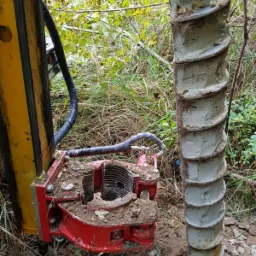 Wiertnica z świdrem w ziemi, widoczne błoto na urządzeniu, w tle trawa i krzewy.