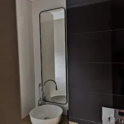 Nowoczesna łazienka z umywalką nablatową na drewnianym blacie, czarnymi płytkami na ścianie i wysokim lustrem w czarnej ramie.