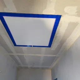 Sufit z płyt kartonowo-gipsowych w trakcie montażu, widoczne łączenia płyt zabezpieczone taśmą, fragment białej wnęki z niebieską taśmą zabezpieczającą.