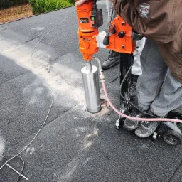 Pracownik wierci otwór w pokryciu dachowym za pomocą wiertnicy diamentowej, widoczne ślady pyłu i przewody