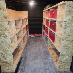 Wnętrze furgonetki z zabudową regałową z płyty OSB, widoczne czerwone skrzynki w przegródkach, oświetlenie sufitowe, podłoga betonowa i narzędzie kątowe przy ścianie.