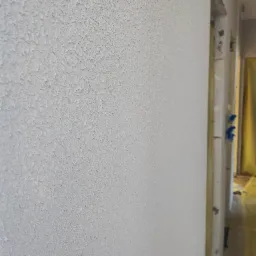 Ściana pokryta jasnoszarą, chropowatą strukturą, z widocznym fragmentem ościeżnicy drzwiowej zabezpieczonej folią malarską w tle.