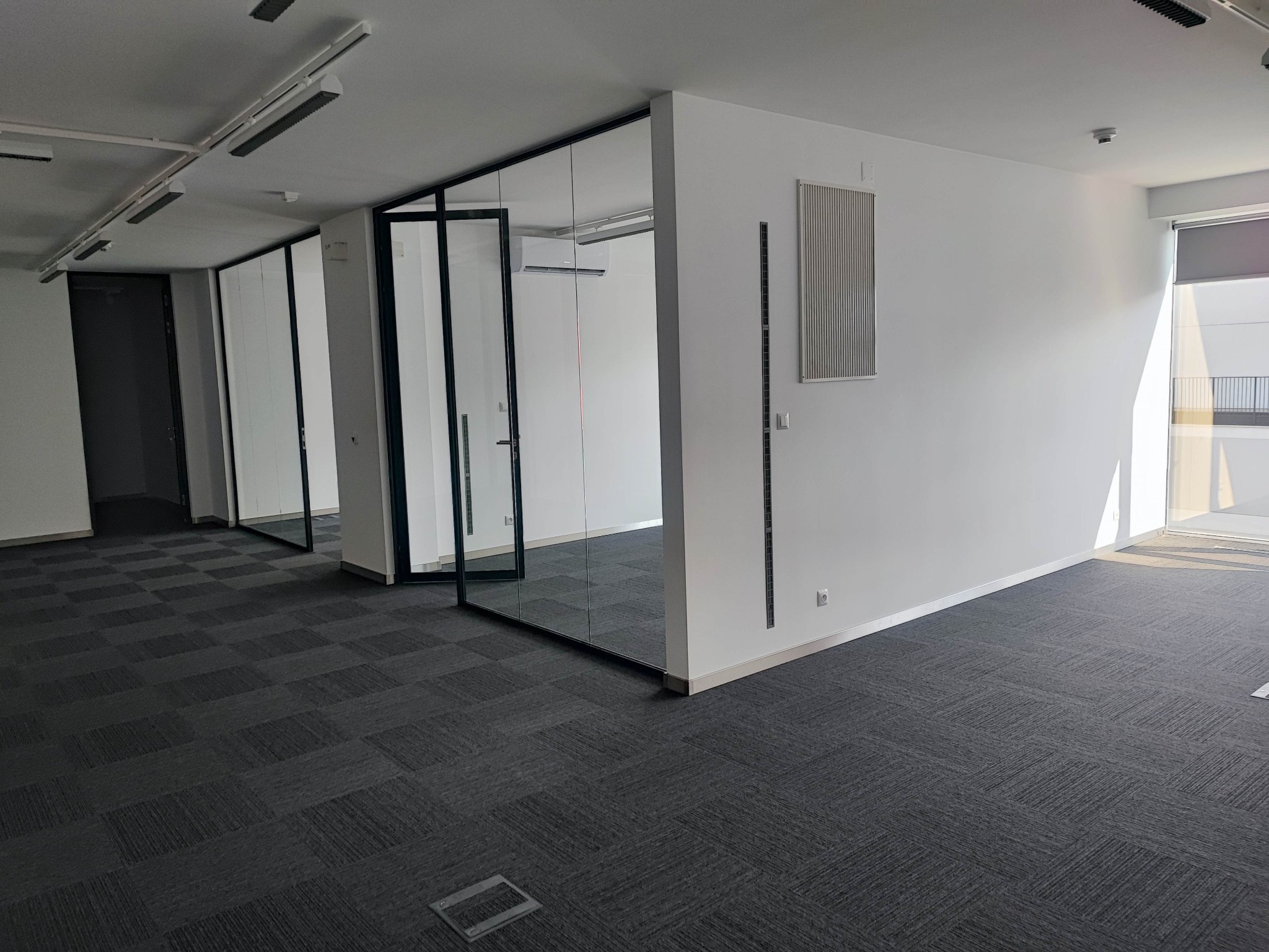 Wykończone biuro z szarą wykładziną dywanową w kwadraty, przeszklonymi ścianami działowymi z czarnymi ramami i białymi ścianami. Widoczny klimatyzator za szkłem i grzejnik na ścianie.