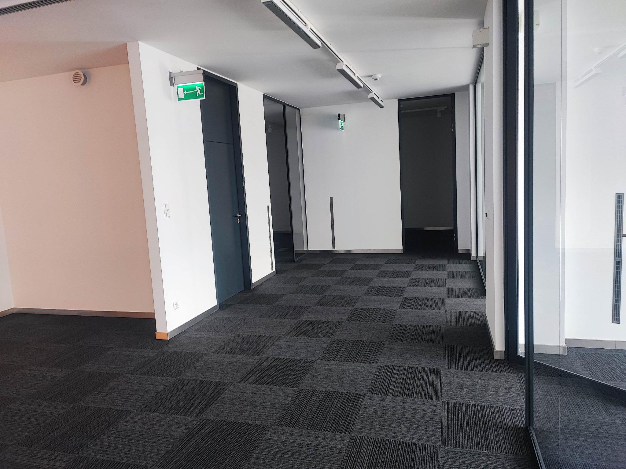 Wnętrze nowoczesnego biura z szarą wykładziną w kwadraty, białymi ścianami, szklanymi drzwiami i oświetleniem liniowym LED oraz zielonym znakiem ewakuacyjnym.