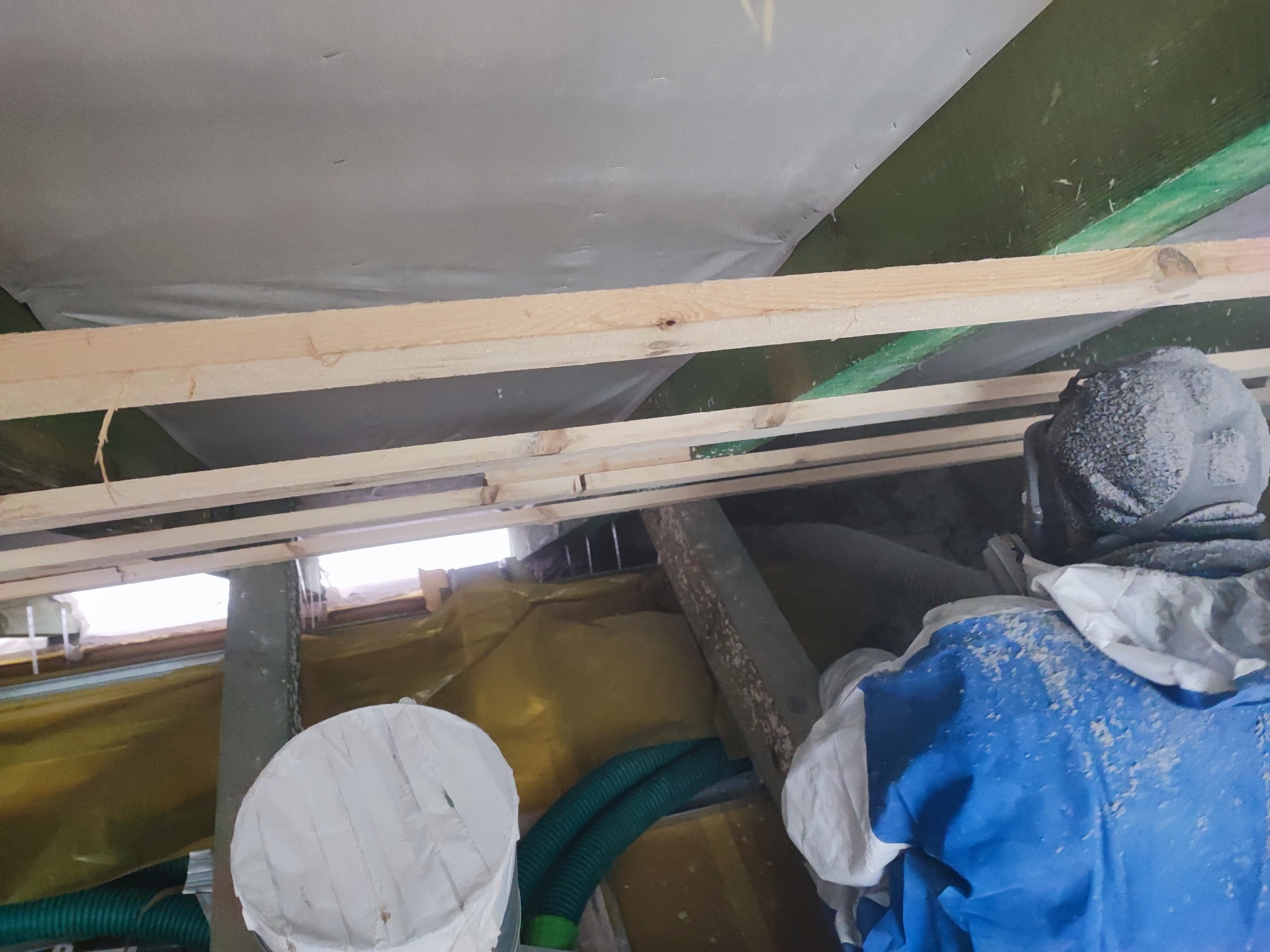 Pracownik w kombinezonie ochronnym i masce podczas izolacji poddasza wełną mineralną, widoczne drewniane belki konstrukcyjne i zabezpieczająca folia. W tle okno.