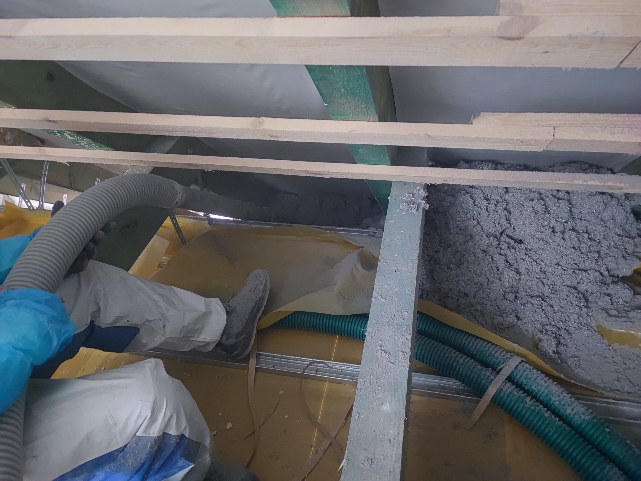 Pracownik w kombinezonie ochronnym aplikuje szarą izolację na poddaszu za pomocą węża do wdmuchiwania, widoczne drewniane belki konstrukcyjne i folia paroizolacyjna.