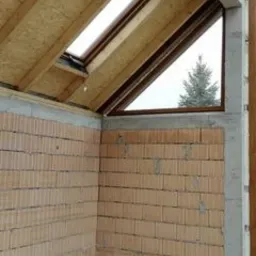 Wnętrze w trakcie budowy z widoczną ścianą z pustaków, betonowymi filarami, drewnianą konstrukcją dachu z oknem połaciowym i trójkątnym oknem z widokiem na drzewo na zewnątrz.