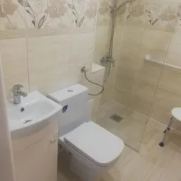 Wykończona łazienka w Dąbrowie Górniczej z beżowymi płytkami, kabiną prysznicową ze szklaną ścianką, umywalką z szafką i toaletą. Widoczny również taboret ułatwiający korzystanie z prysznica.