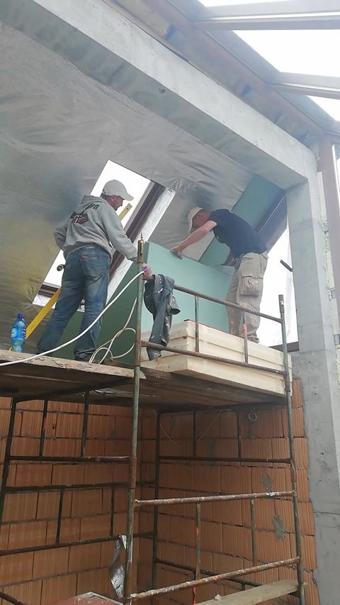 Dwóch pracowników na rusztowaniu montuje zielone płyty na poddaszu, widoczna izolacja termiczna i ceglana ściana.