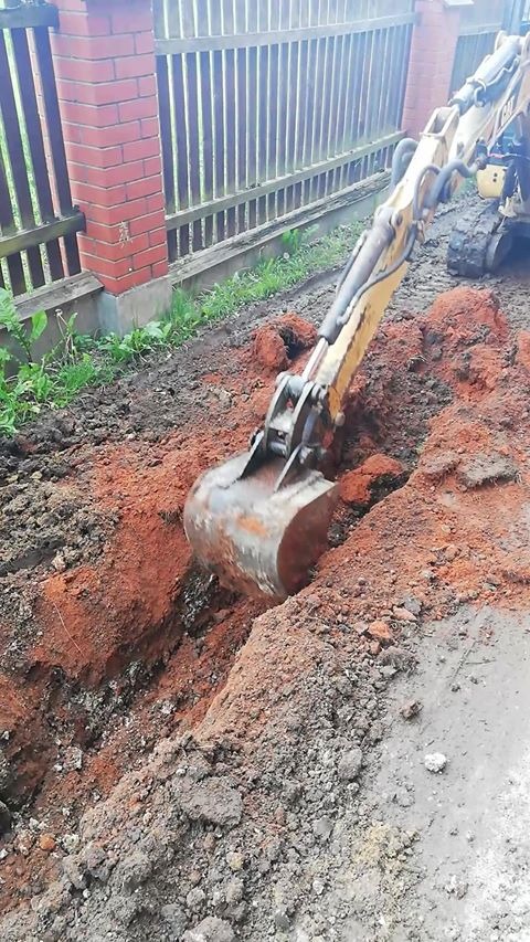 Żółta koparka wykopuje wąski rów w czerwonej ziemi wzdłuż drewnianego płotu z ceglanym słupkiem.