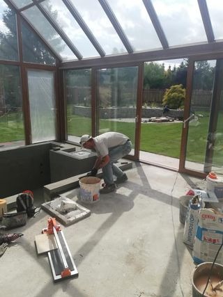 Praca glazurnicza wewnątrz przeszklonej werandy: mężczyzna w czapce układa płytki na betonowej konstrukcji, widoczne narzędzia, wiadro z zaprawą, torby z klejem. Tło to zielony ogród widziany przez...
