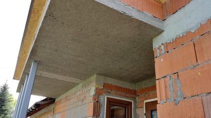 Surowy stan elewacji budynku z widoczną warstwą izolacyjną nad oknem, ceglaną ścianą i elementami konstrukcyjnymi z betonu.