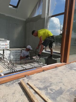 Dwóch pracowników montuje zbrojenie w nowo budowanym pomieszczeniu z dużym oknem i świetlikiem dachowym, widoczne materiały izolacyjne i surowe ściany.