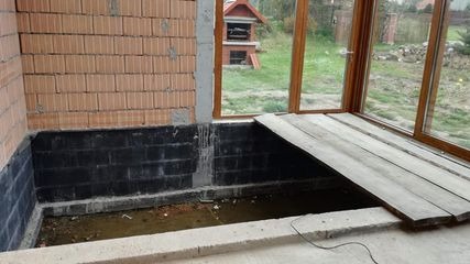 Niedokończona wylewka fundamentu z hydroizolacją pionową na ścianie, widoczne cegły, okno balkonowe i deski pomostowe