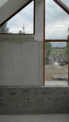 Surowa ściana wewnętrzna z niedokończonymi gładziami, widoczne okna dachowe z drewnianymi ramami i widokiem na zewnętrzny teren budowy, otwory na instalacje w dolnej części ściany.