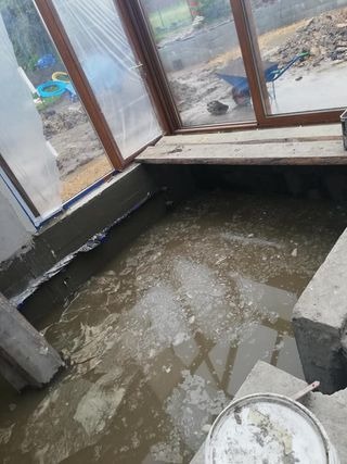 Wnętrze balkonu w trakcie prac remontowych, zalane wodą z resztkami materiałów budowlanych, widoczne okna balkonowe z drewnianą ramą i zabezpieczone folią. W tle widoczny teren budowy.