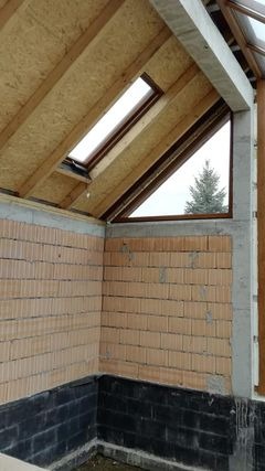 Wnętrze w trakcie budowy z widoczną ścianą z pustaków, betonowymi filarami, drewnianą konstrukcją dachu z oknem połaciowym i trójkątnym oknem z widokiem na drzewo na zewnątrz.