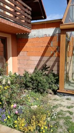 Fragment ściany budynku z pustaków ceramicznych, częściowo otynkowany, z widocznym ogrodem i elementami konstrukcji dachu