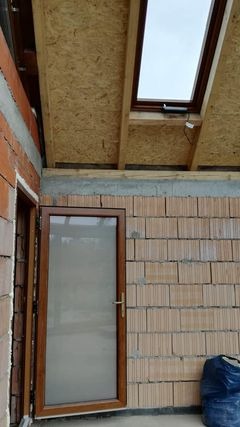 Wnętrze budynku w trakcie budowy z widoczną ścianą z cegieł, drewnianą ramą okienną w suficie i częściowo zamontowanymi drzwiami z brązową ramą.