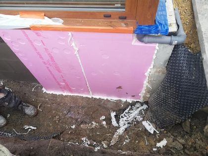 Różowa płyta termoizolacyjna XPS przyklejona do fundamentu budynku, uszczelniona pianką montażową, widoczna folia kubełkowa i rura odpływowa.