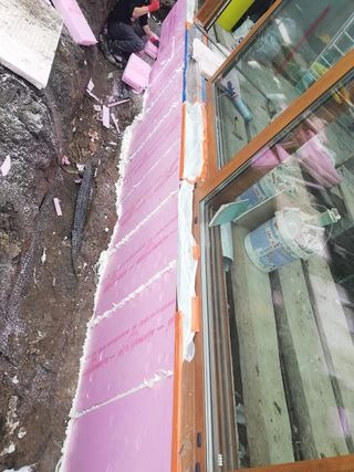 Ocieplanie elewacji różowym styropianem przy oknie, widoczna warstwa kleju i pracownik w trakcie montażu.