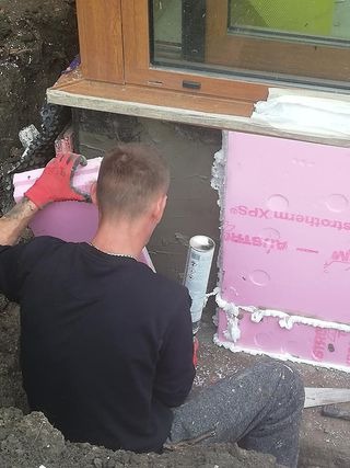 Mężczyzna w czerwonych rękawicach aplikuje piankę montażową między różowe płyty izolacyjne Austrotherm XPS przy fundamencie budynku, obok okna.