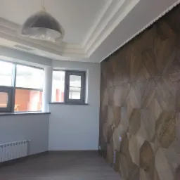 Pokój z drewnianą podłogą i ścianą z paneli 3D w odcieniach brązu, okna z szarymi ramami, biały sufit z dekoracyjnymi listwami, lampa wisząca zabezpieczona folią.