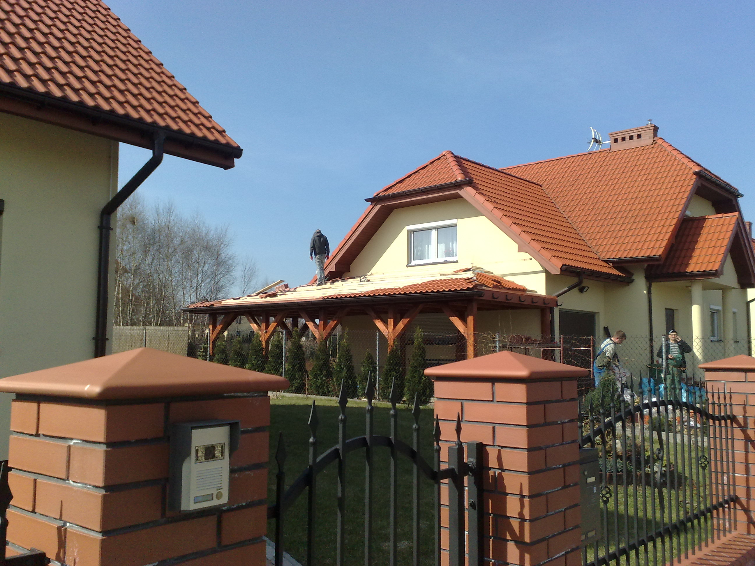 Budowa domu jednorodzinnego z czerwonym dachem: dekarz pracuje na drewnianej konstrukcji dachu, widoczny fragment elewacji, ogrodzenie z ceglanymi słupkami i metalową furtką.
