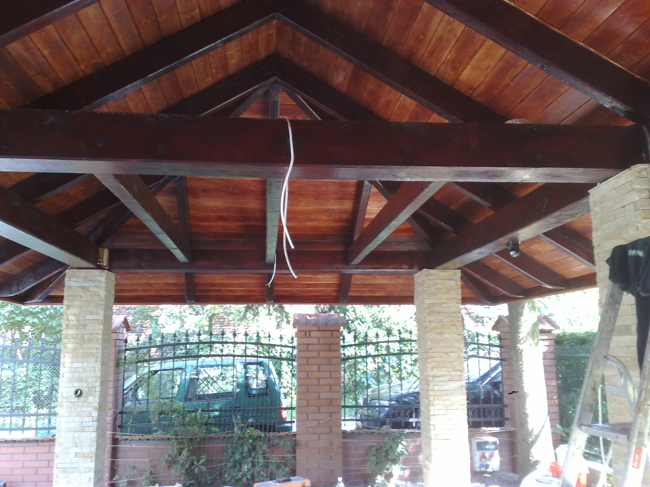 Drewniana konstrukcja zadaszenia z widocznymi belkami i deskami, wsparta na murowanych filarach, z widokiem na ogrodzenie i pojazdy w tle.