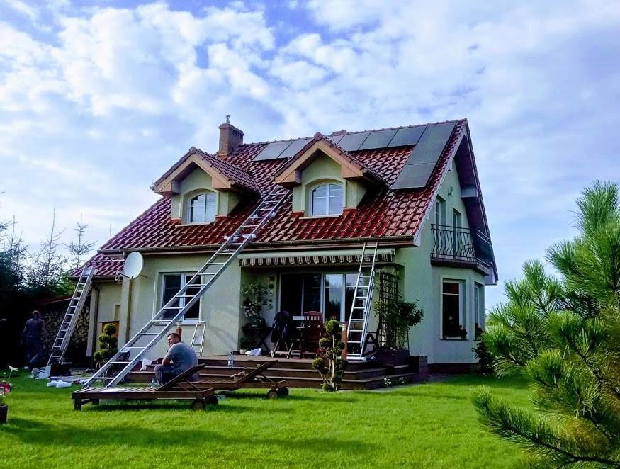 Dom jednorodzinny z panelami fotowoltaicznymi na dachu, ustawione drabiny i osoba siedząca na leżaku, prawdopodobnie podczas przerwy w montażu.