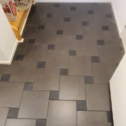 Ułożona podłoga z brązowych, kwadratowych płytek z ciemnoszarymi wstawkami, widoczne pudełka z gaśnicami w tle.