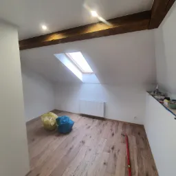 Poddasze po remoncie: drewniane belki stropowe, okno dachowe, białe ściany, panele podłogowe, grzejnik, worki z odpadami i narzędzia malarskie na półce.