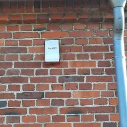 Zewnętrzna obudowa alarmowa firmy AL-CAM zamontowana na ceglanej ścianie budynku, widoczna rynna spustowa po prawej stronie.