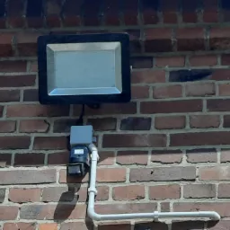 Zewnętrzny reflektor LED z czujnikiem ruchu zamontowany na ceglanej ścianie, widoczne przewody i rurki instalacyjne.