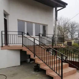 Zewnętrzne schody wejściowe do domu, wyłożone terakotą, z czarną, metalową balustradą po obu stronach, widok na elewację budynku z oknami.