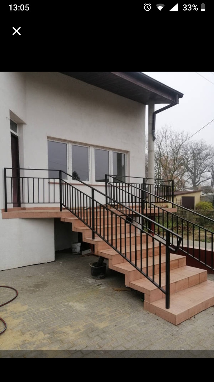 Zewnętrzne schody wejściowe do domu, wyłożone terakotą, z czarną, metalową balustradą po obu stronach, widok na elewację budynku z oknami.