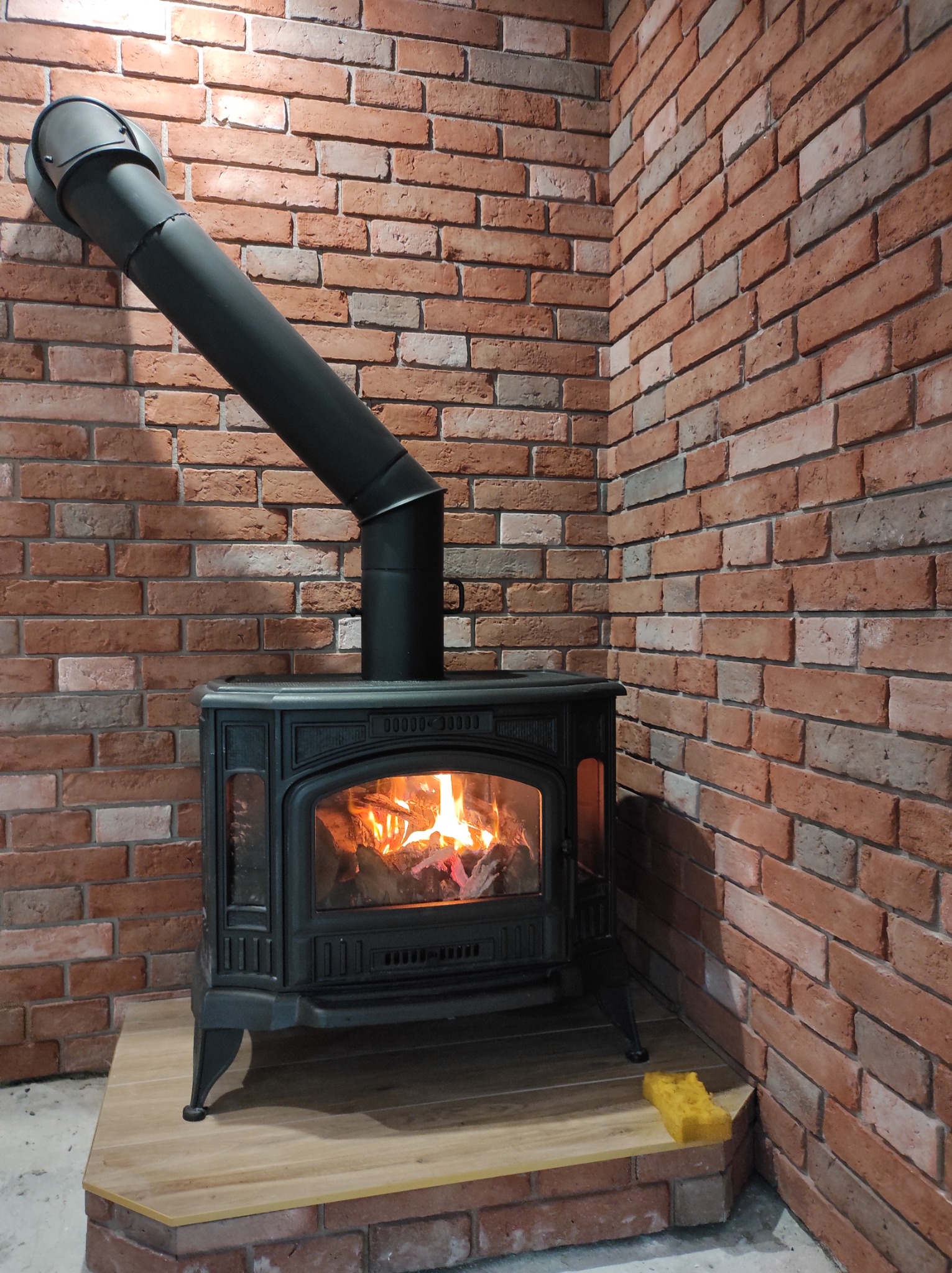Żeliwny piec kominkowy z rurą odprowadzającą dym, umieszczony na drewnianym podeście wykończonym cegłą dekoracyjną, płomienie widoczne za szybą, z boku leży żółta gąbka.