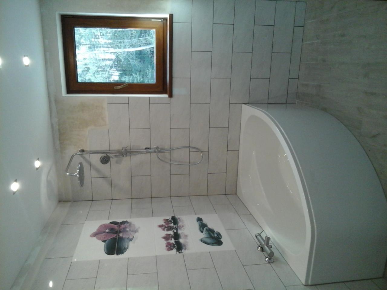 Biała, narożna wanna w łazience wyłożonej jasnymi, prostokątnymi płytkami; na podłodze płytki z motywem kwiatowym, widoczna armatura i okno z drewnianą ramą.