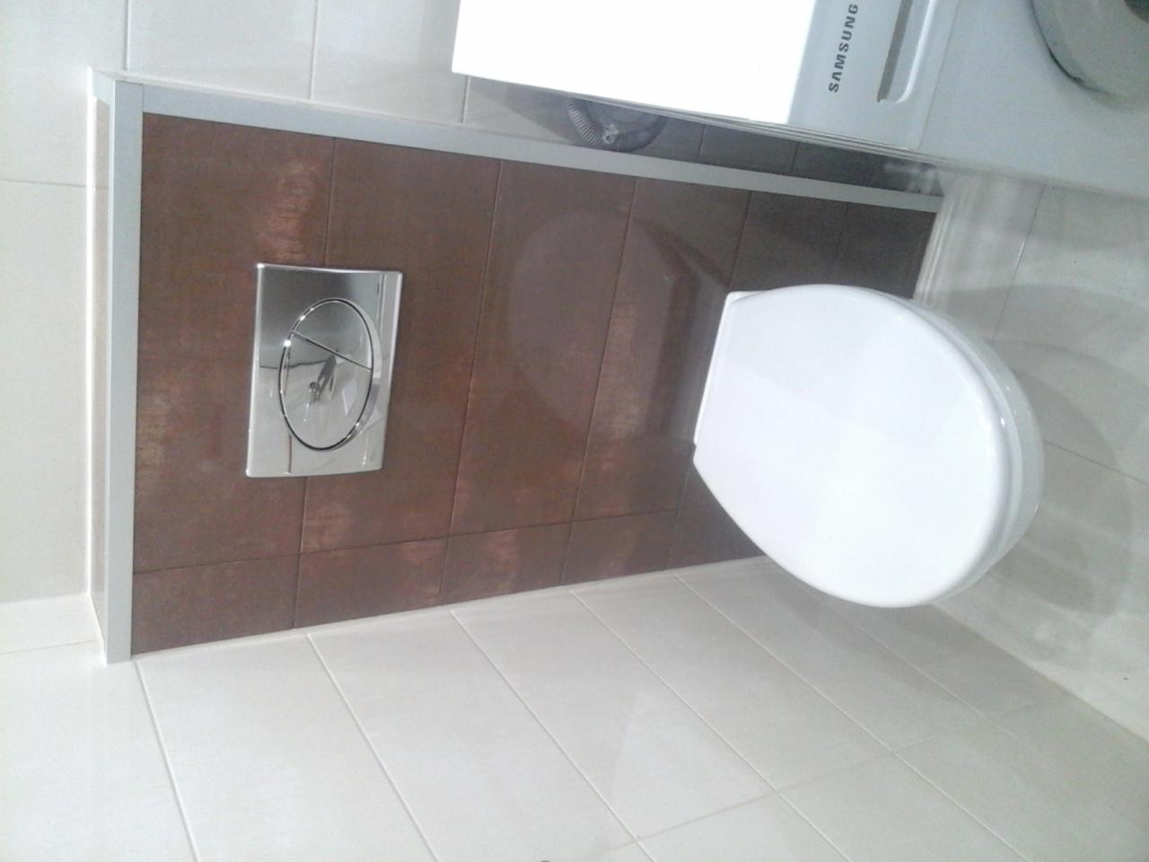 Toaleta podwieszana z brązowymi płytkami ściennymi i chromowanym przyciskiem spłukiwania, widoczna z góry, z fragmentem pralki Samsung w tle.