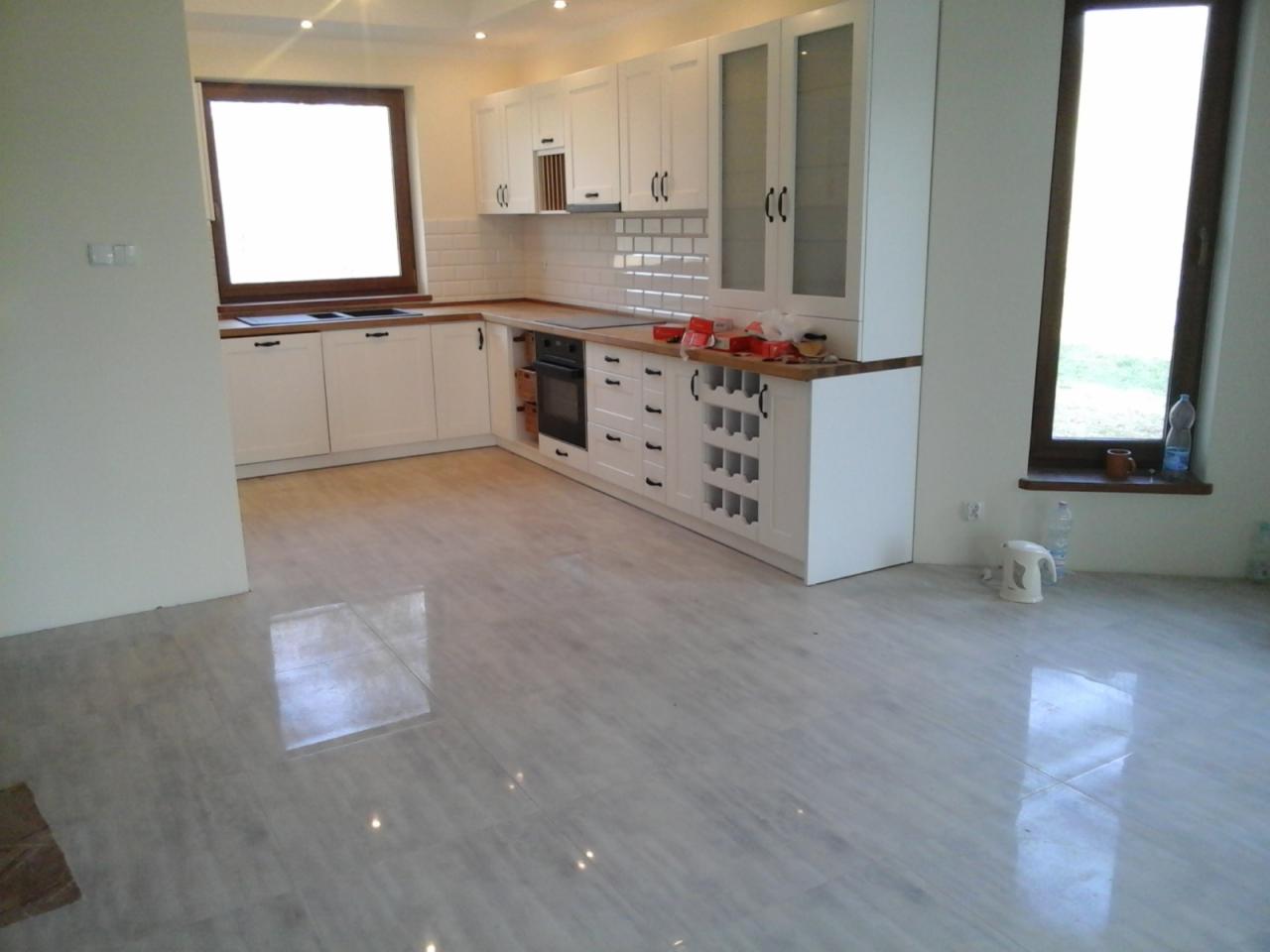 Jasna kuchnia z białymi szafkami i drewnianymi blatami, widok na błyszczące, szare płytki podłogowe i okno z widokiem na zieleń, czajnik elektryczny i butelka wody na parapecie.