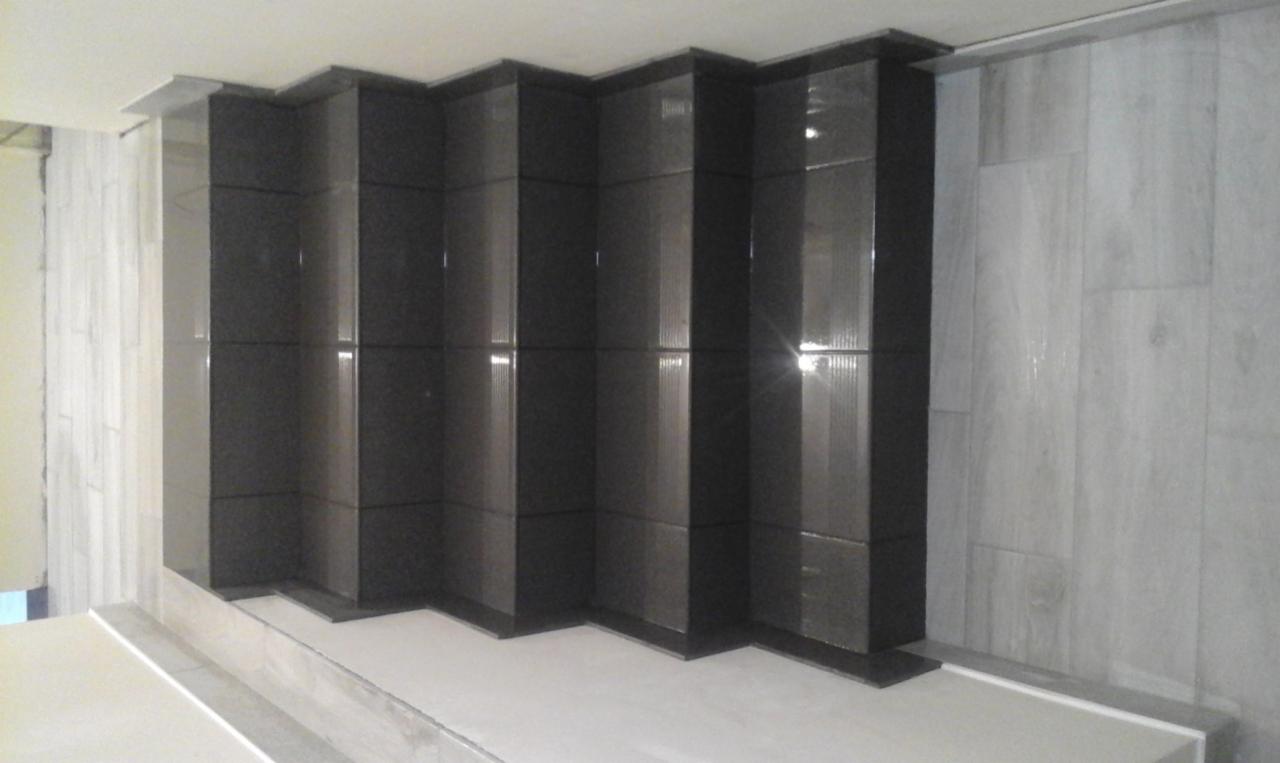 Ściana wyłożona naprzemiennie czarnymi błyszczącymi płytkami tworzącymi efekt schodkowy oraz jasnymi płytkami imitującymi drewno.